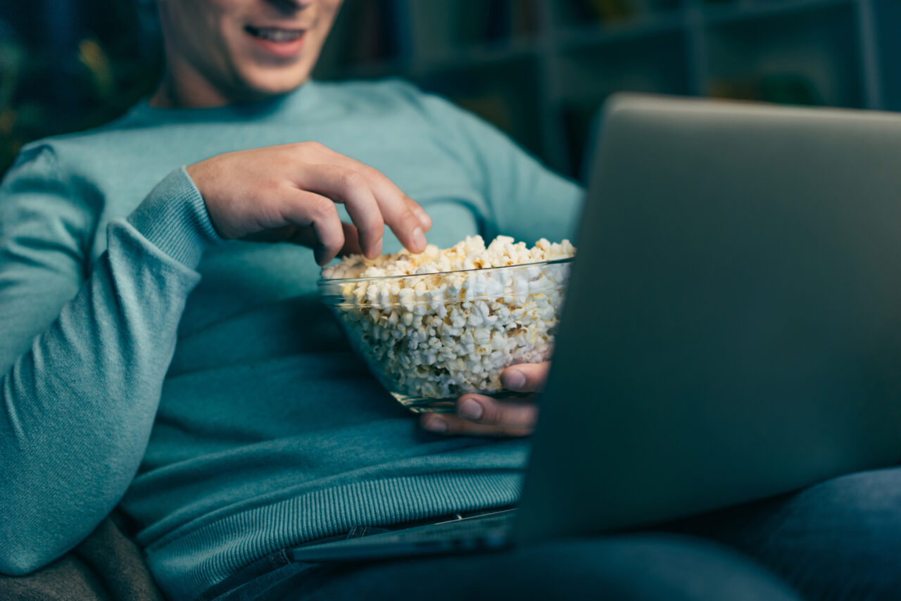 Mężczyzna siedzący na kanapie przed laptopem bierze garść popcornu z przezroczystej miski.
