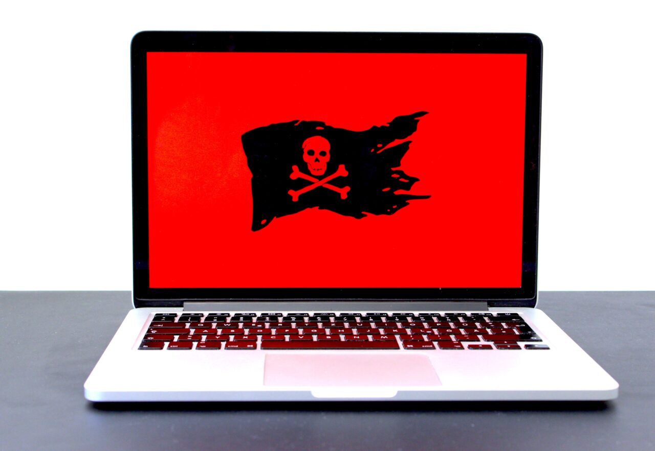 Laptop na biurku z wyświetlonym na ekranie obrazem pirackiej flagi - czarnej czaszki i skrzyżowanych kości na czerwonym tle.