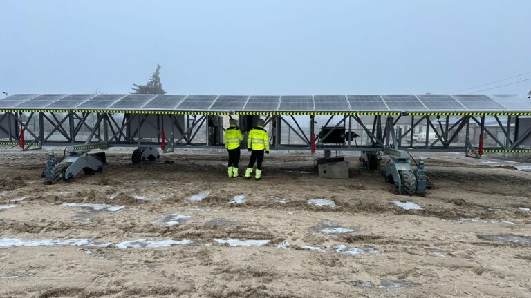 Widok dwóch osób w odblaskowych kamizelkach stojących na budowie farmy fotowoltaicznej, z zainstalowanymi panelami słonecznymi i rozpościerającymi się przewodami ostrzegawczymi, na tle zimowego, lekko zaśnieżonego krajobrazu.