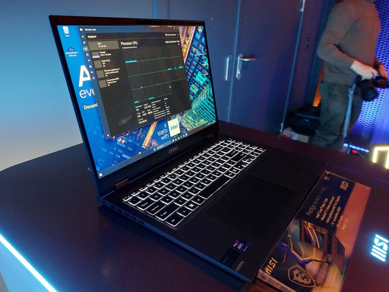 Laptop firmy MSI ustawiony na biurku z włączonym ekranem pokazującym otwarte okno monitora wydajności. W tle częściowo widoczny jest mężczyzna oraz neonowe oświetlenie w pokoju.