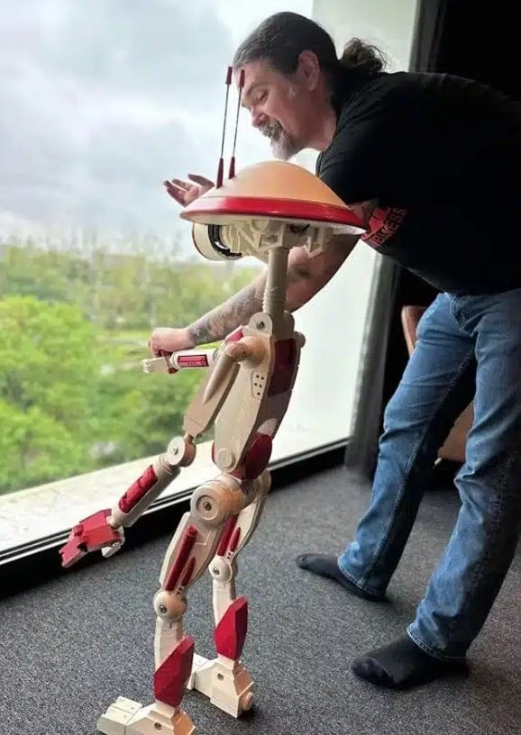 Mężczyzna obserwuje drewnianego robota na cięciwach, który jest podnoszony. Robot przypomina postać humanoida i stoi obok okna z widokiem na zielone tereny.