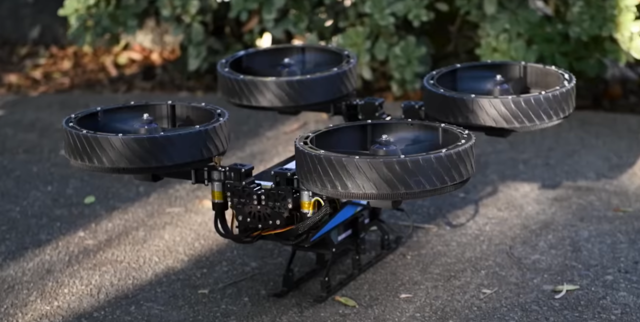 Czterowirnikowy dron z czarnymi osłonami śmigieł, stojący na asfaltowej ścieżce obok krzewów.