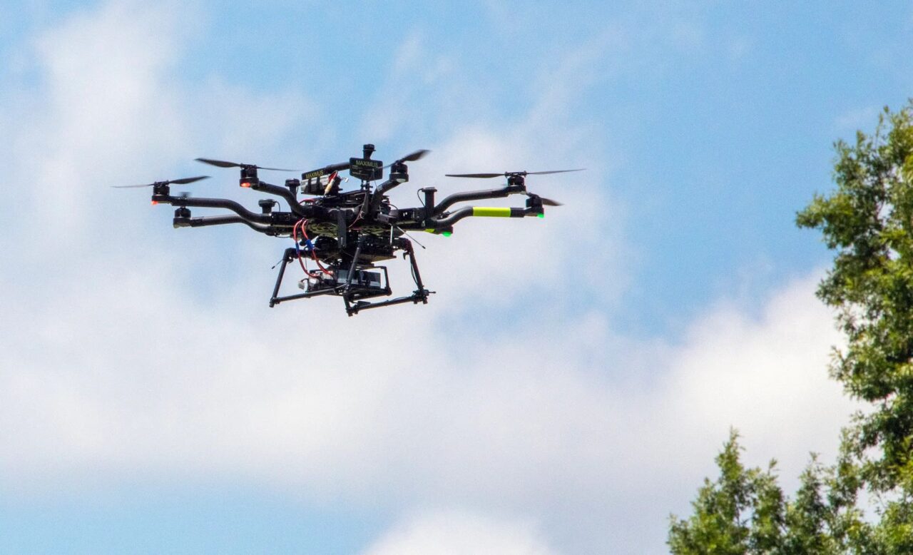 Profesjonalny wielowirnikowy dron NASA latający na niebieskim niebie z chmurami i drzewami w tle.
