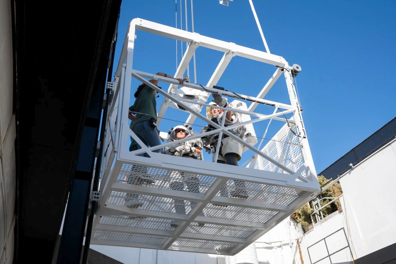 Podnoszona na dźwigu platforma NASA z ludźmi w kaskach i ubraniach roboczych.