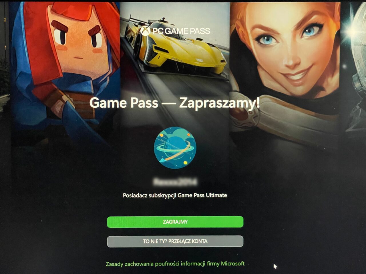 Ekran komputera z grafiką reklamującą usługę Xbox Game Pass z postaciami z gier, samochodem wyścigowym i napisem "Game Pass — Zapraszamy!". Na dole widoczne przyciski "ZAGRAJMY" oraz "TO NIE TY? PRZEŁĄCZ KONTA" oraz logo firmowe.