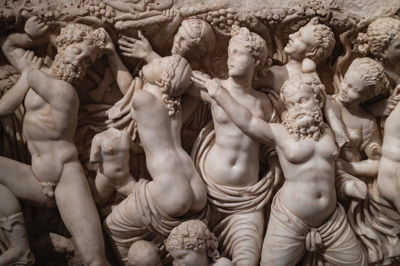 Detal wysokiej jakości rzeźby w marmurze przedstawiającej postacie mitologiczne w dynamicznych pozach z wyraźnie zarysowanymi detalami i drobnymi akcentami jak girlandy i tkaniny.