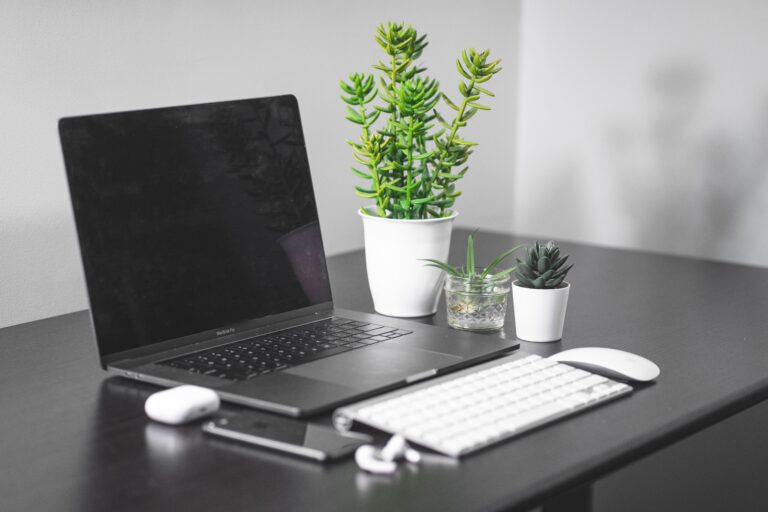 Zdjęcie biurka z otwartym laptopem, obok leżą słuchawki i telefon, a za urządzeniami stoją doniczki z zielonymi roślinami.