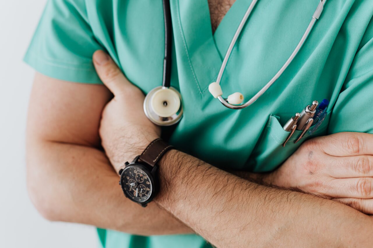 Mężczyzna w zielonym uniformie medycznym z założonymi rękami, na którego nadgarstku znajduje się zegarek, a w kieszeni uniformu przymocowany jest stetoskop i długopis.