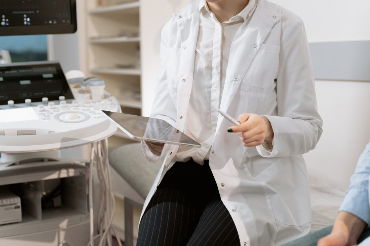 Osoba w białym lab-coat trzymająca kartę rentgenowską i długopis w gabinecie medycznym, z urządzeniem ultrasonograficznym w tle. Może na tablecie pokazywać e-recepty lub obsługiwać e-ZLA z ZUS.