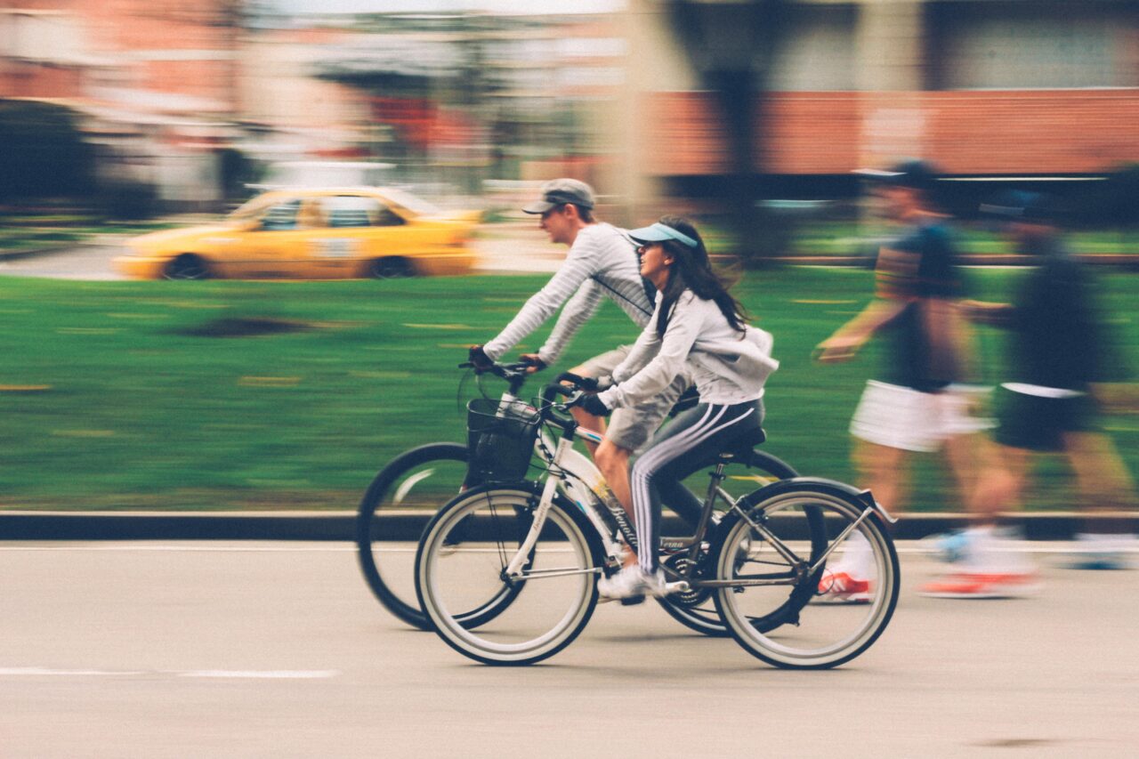 W co powinien wyposażyć się nowoczesny rowerzysta? Oto najlepsze gadżety na rower. Dwóch rowerzystów w ruchu na tle nieostrego żółtego taksówki i pieszych, zdjęcie o rozmytym tle, podkreślającym szybkość.