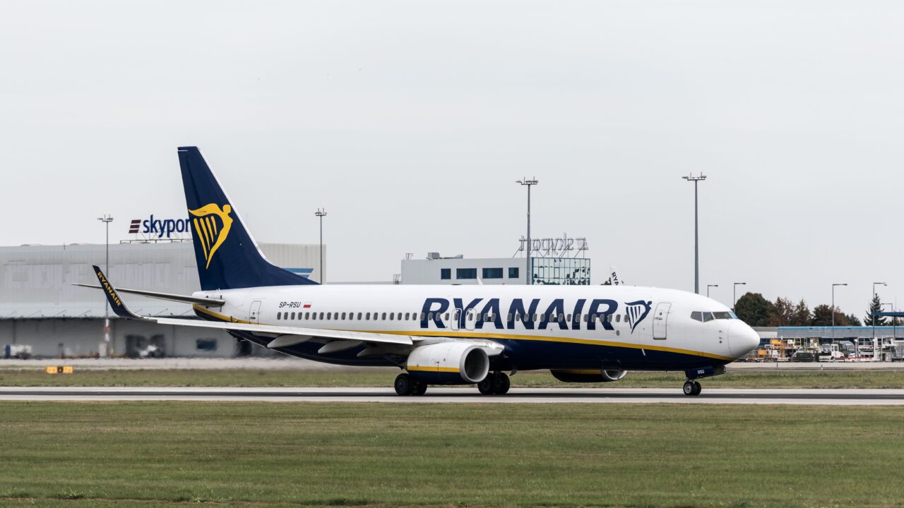 Samolot pasażerski Ryanair na pasie startowym z budynkami lotniskowymi w tle.