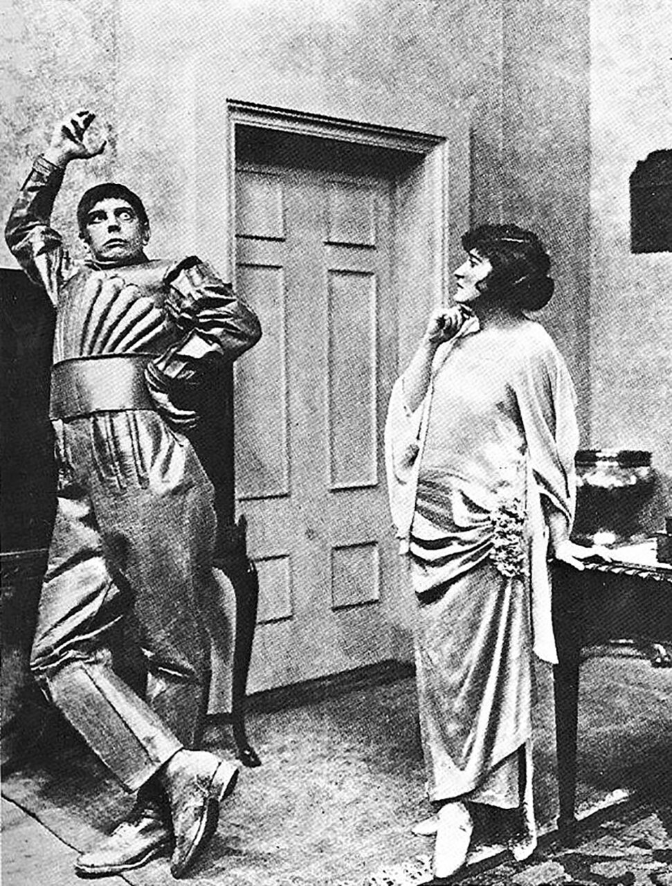 Czarno-białe zdjęcie przedstawiające mężczyznę w pasiastym kostiumie, który wygląda przestraszony, stojącego obok kobiety w długiej sukni, która wydaje się być zaniepokojona. Scena rozgrywa się w pomieszczeniu z drzwiami w tle.