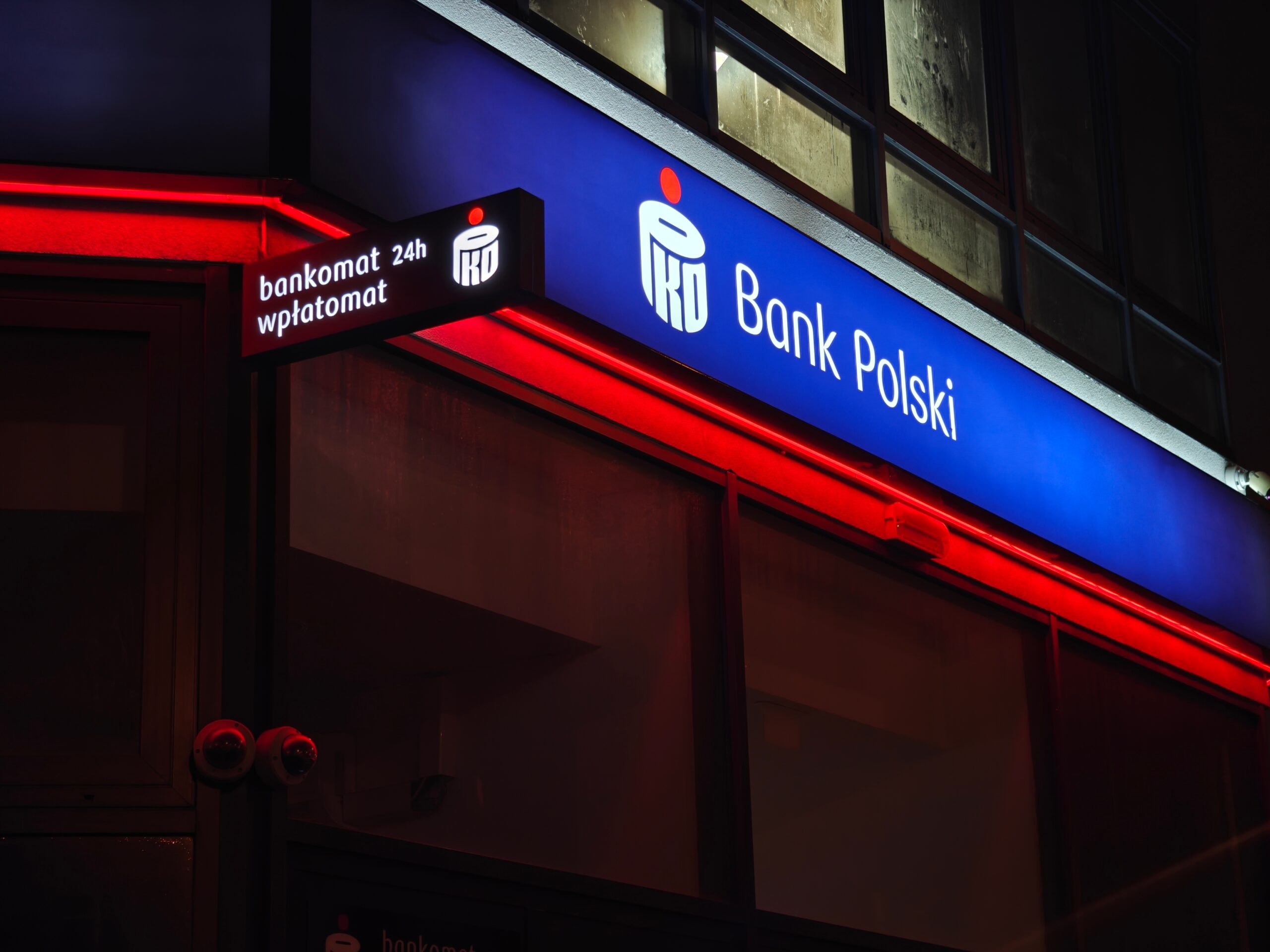 Zdjęcie neonowego szyldu PKO Bank Polski i znaku informacyjnego z napisem "bankomat 24h wpłatomat" w nocy.