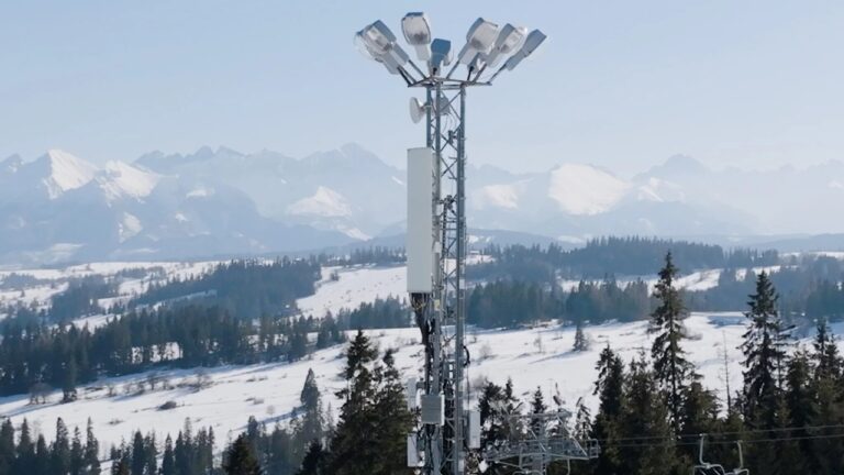 Maszt z urządzeniami telekomunikacyjnymi i oświetleniem na tle zaśnieżonych gór i lasów.