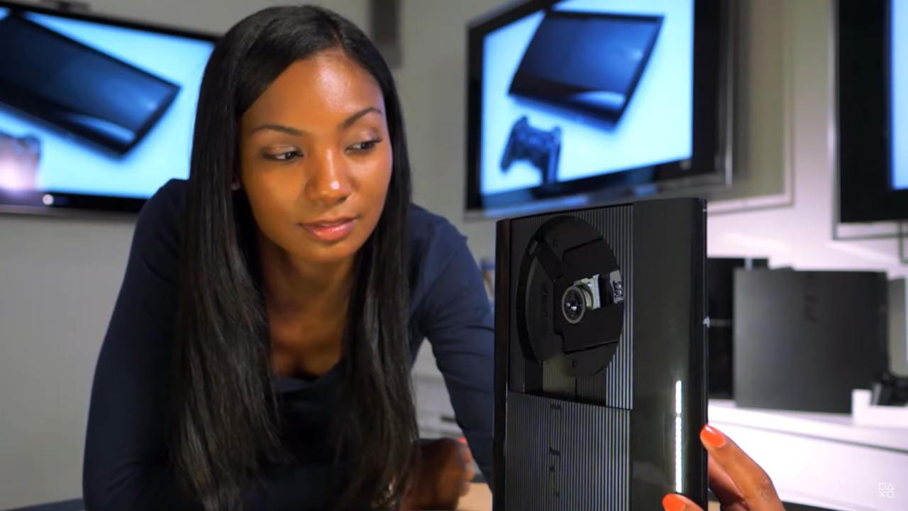 Kobieta patrzy na otwartą konsolę do gier PlayStation 3, która ujawnia wewnętrzne komponenty, w tle monitory z obrazem konsoli i kontrolera.