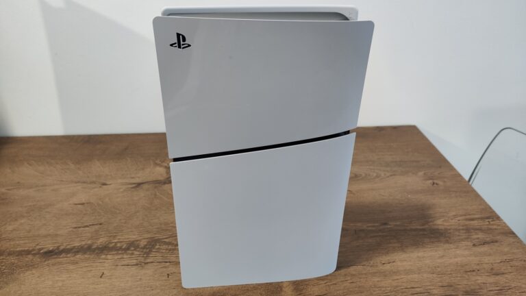 Konsola do gier PlayStation 5 Slim stojąca na drewnianym biurku obok białej ściany.