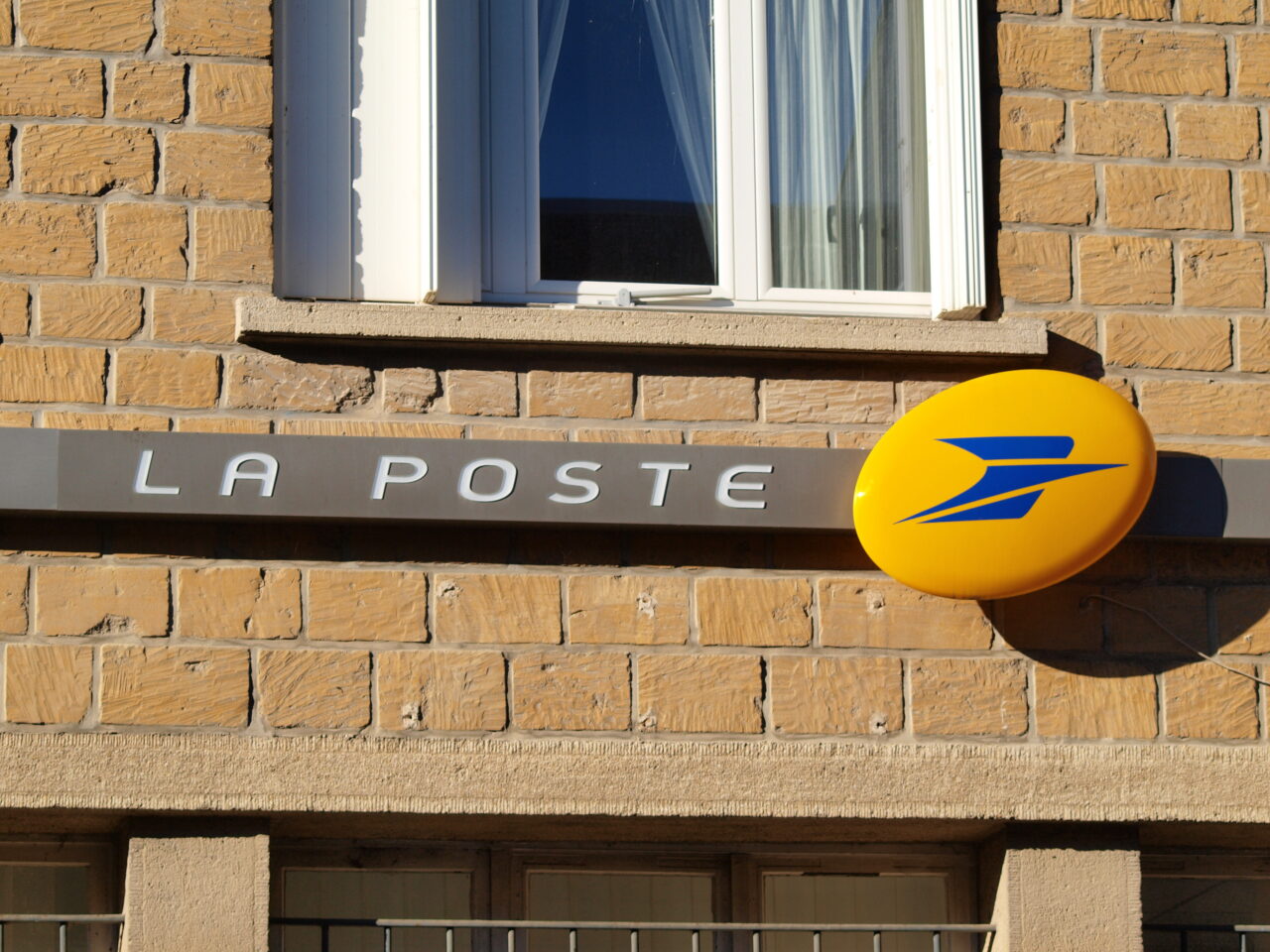 Zdjęcie ściany budynku z oknem oraz znakiem poczty "LA POSTE" wraz z okrągłym, żółtym logo.