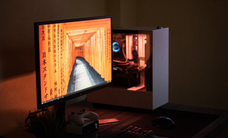 Zdjęcie dobrze oświetlonego domowego stanowiska komputerowego z monitorem, na którym wyświetlany jest obraz pomarańczowego korytarza w chramie Fushimi Inari Taisha w Japonii, obok biała obudowa komputera z podświetleniem, klawiatura, mysz komputerowa i biały kontroler do gier.