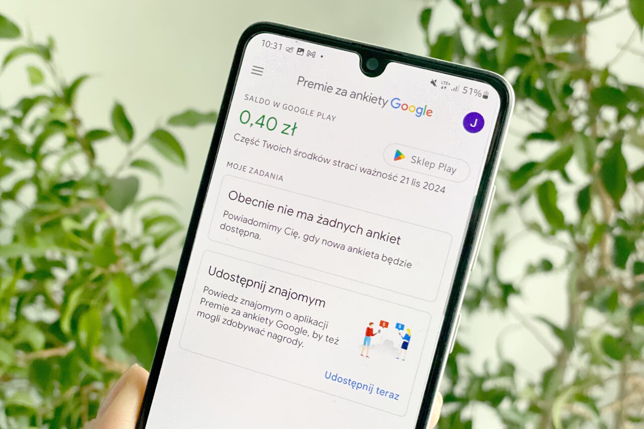 Telefon komórkowy wyświetlający ekran aplikacji "Premie za ankiety Google" z widocznym saldem 0,40 zł oraz komunikatami na tle zielonych roślin.