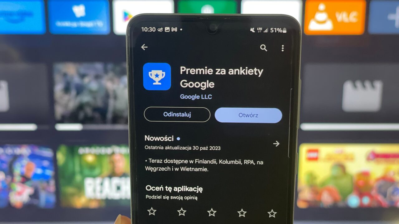 Ręka trzymająca smartfon z otwartą aplikacją Google Opinion Rewards na ekranie, z tłem przedstawiającym rozmyte ikony aplikacji na ekranie telewizora.