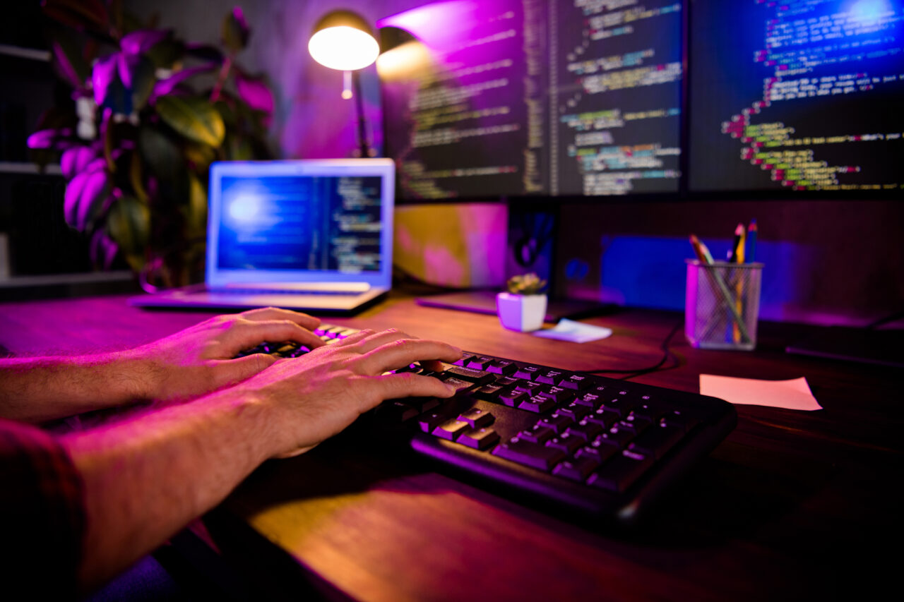 Ręce programisty piszącego kod na klawiaturze w mrocznym biurze z podświetleniem, z widocznym w tle laptopem i monitorem z kodem programistycznym.