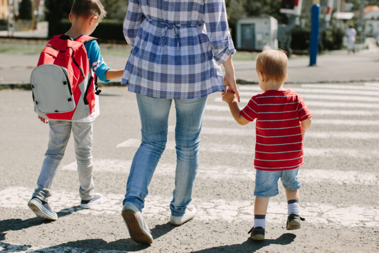Kobieta przechodzi przez przejście dla pieszych trzymając za rękę małego chłopca w czerwonej koszulce w paski, a drugi chłopiec z plecakiem idzie obok.