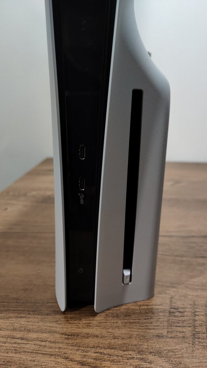 Konsola do gier PlayStation 5 Slim stojąca pionowo na drewnianym blacie, z widocznymi portami USB i przyciskiem zasilania.