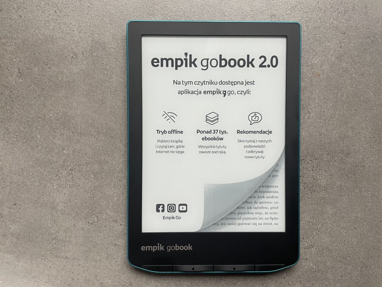 Czytnik e-booków Empik GoBook 2.0 leżący na szarym tle, z wyświetlonym ekranem głównym, na którym widnieje informacja o dostępnej aplikacji Empik Go.