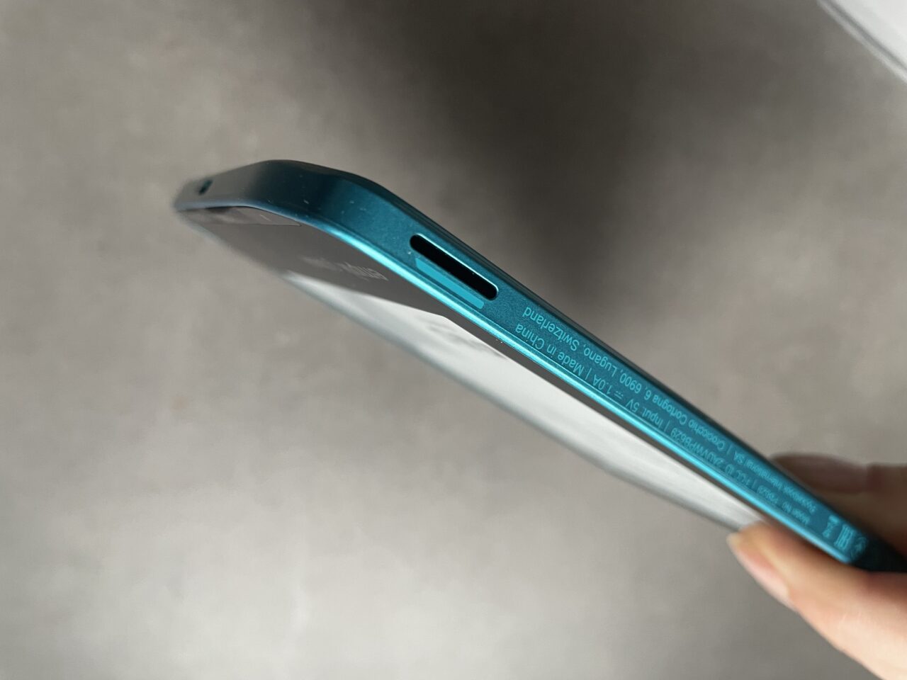 Niebieski smartfon trzymany w dłoni, ukazany skośnie od góry, z widocznym włącznikiem oraz slotem na kartę SIM lub microSD.
