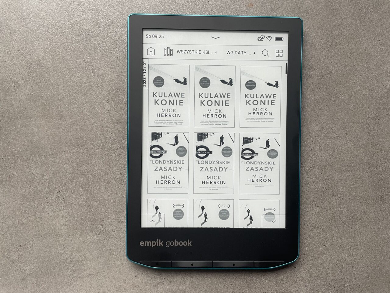 Czytnik e-booków leżący na szarym tle, wyświetlający bibliotekę książek elektronicznych z okładkami autorstwa Micka Herrona, w tym powtórzenia "Kulawe konie" i "Londyńskie zasady". Czytnik ma zaznaczoną ramkę w kolorze turkusowym i logo "empik goBook" na dole.