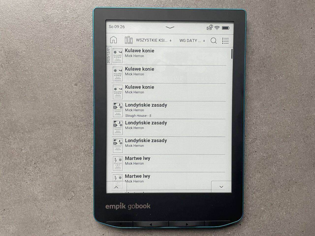 Czytnik ebooków z otwartą listą tytułów książek na ekranie, na szarym tle.