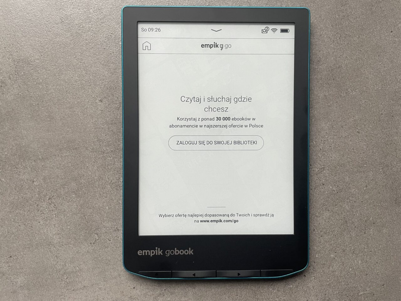Czytnik ebooków leżący na szarym tle wyświetla ekran startowy z reklamą usługi empik go.