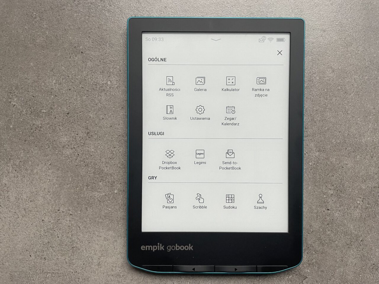 Czytnik e-booków leżący na szarym tle, wyświetlający ekran główny z ikonami aplikacji i funkcji takimi jak galeria, kalkulator i ustawienia.