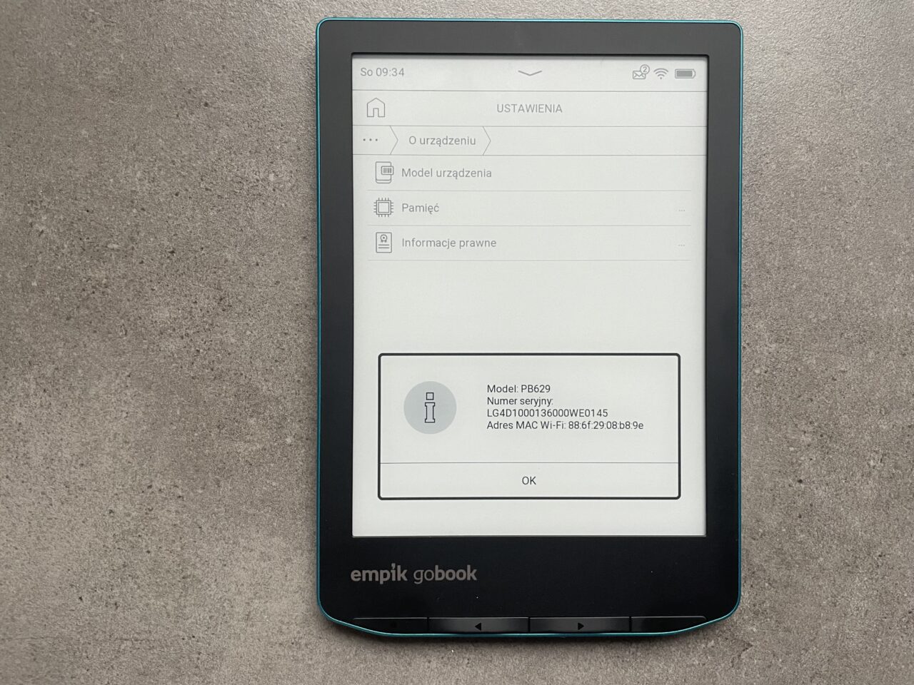 Czytnik ebooków leżący na szarym blacie z wyświetlonym menu ustawień i informacjami o urządzeniu na ekranie.