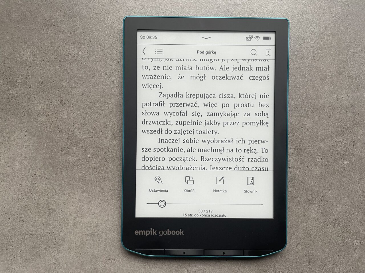Czytnik e-booków leżący na szarym tle, wyświetlający tekst książki z zaznaczonym interfejsem użytkownika.