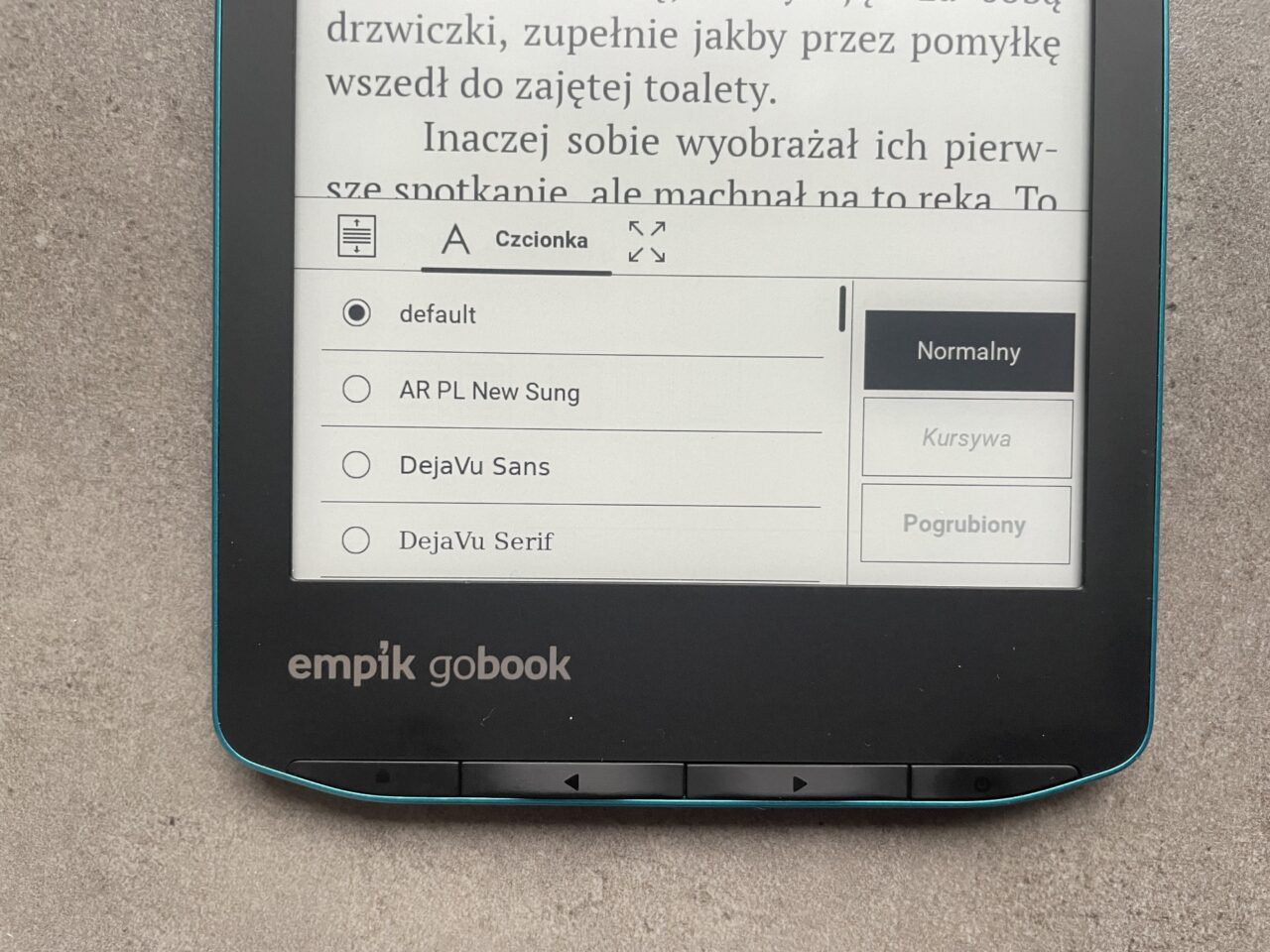 Czytnik e-booków Empik GoBook wyświetlający menu ustawień czcionki z zaznaczoną opcją "default" oraz opcjami formatowania tekstu "Normalny", "Kursywa", i "Pogrubiony" obok fragmentu otwartej książki elektronicznej.