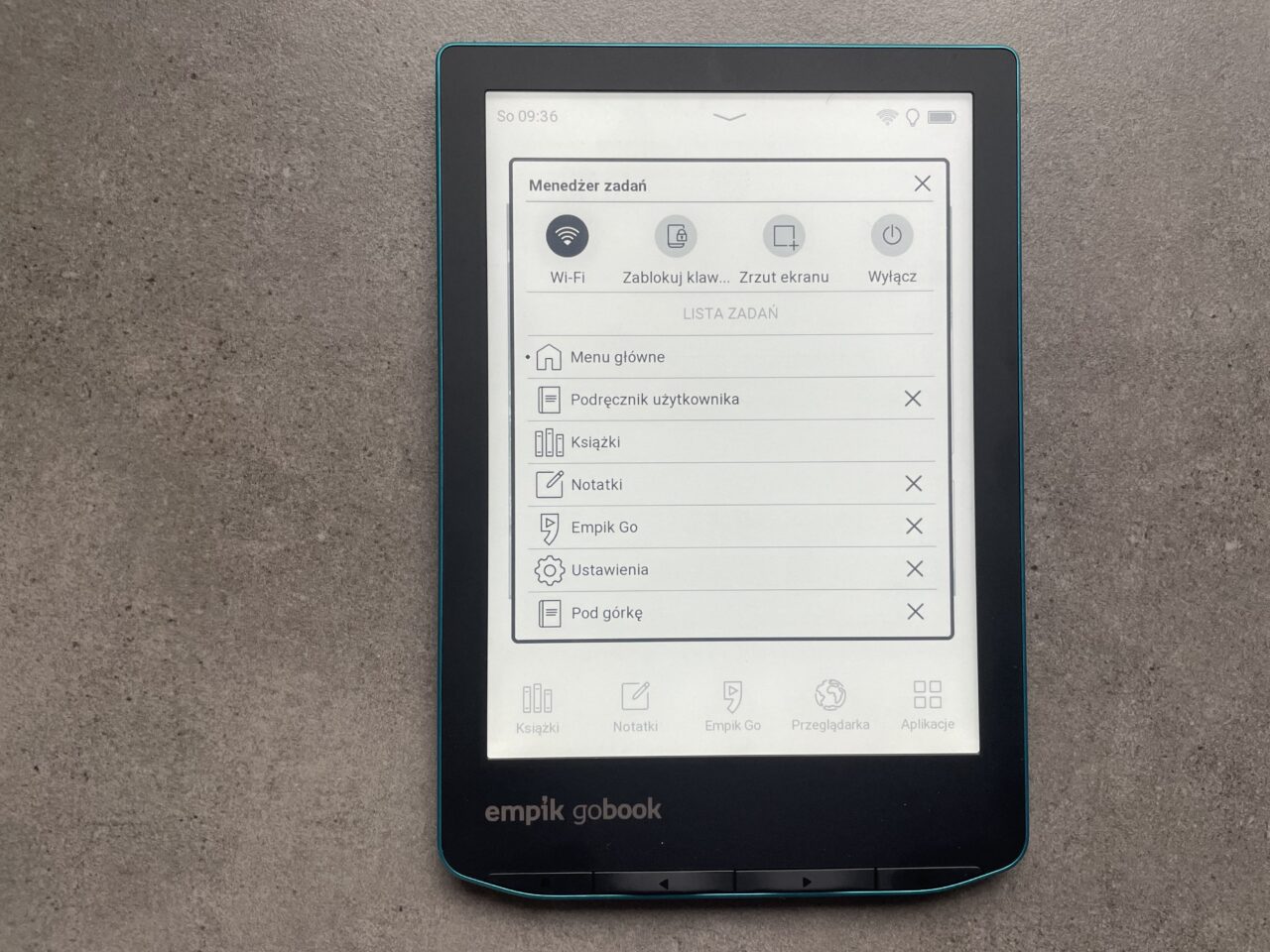 Czytnik e-booków leżący na szarym tle z otwartym ekranem menu z opcjami takimi jak Wi-Fi, Lista zadań, Ustawienia i ikonami aplikacji na dole ekranu.