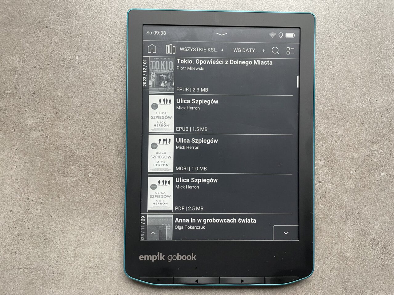 Czytnik e-booków leżący na szarym tle z wyświetloną listą książek w bibliotece cyfrowej.