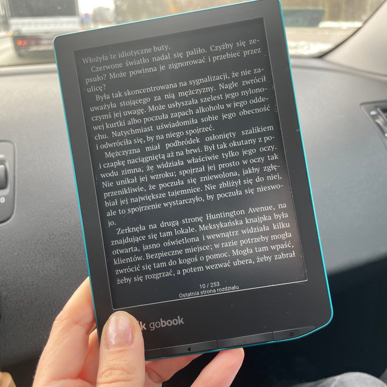 Osoba trzyma czytnik ebooków z włączonym tekstem, w tle widoczne jest wnętrze samochodu oraz okno z rozmytym obrazem jadących pojazdów.
