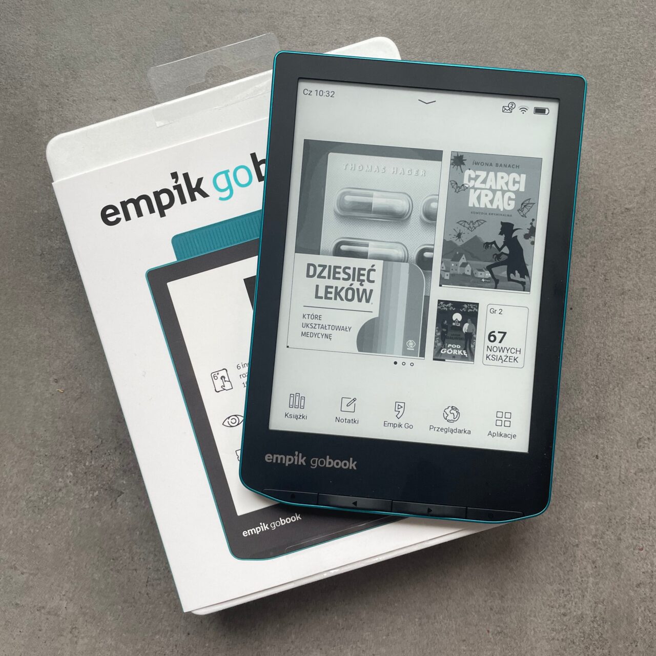 Czytnik e-booków Empik GoBook na pudle, z włączonym ekranem wyświetlającym interfejs z okładkami książek, leżący na szarym tle.