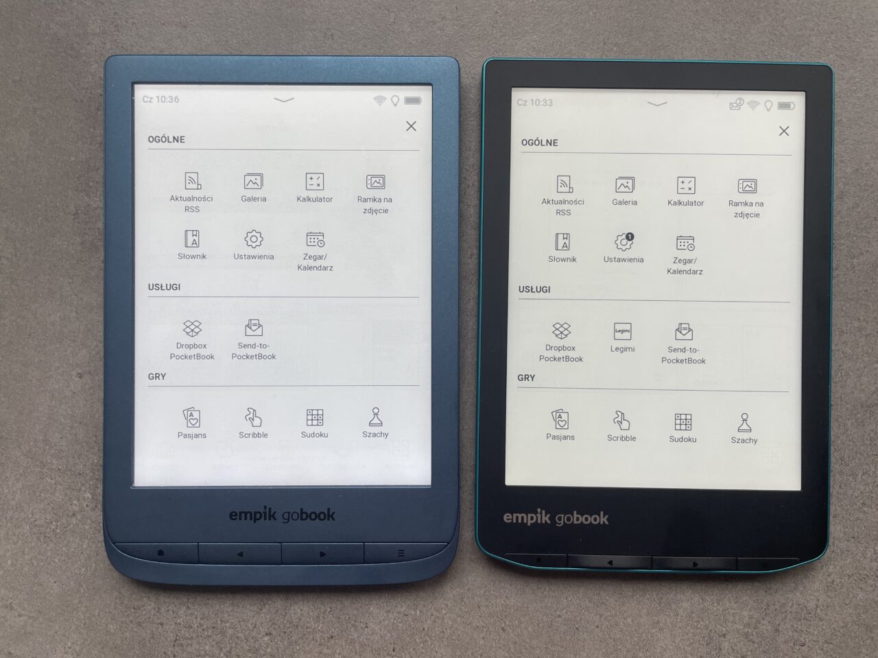 Dwa czytniki ebooków Empik GoBook z włączonymi ekranami głównego menu, leżące obok siebie na szarym tle.