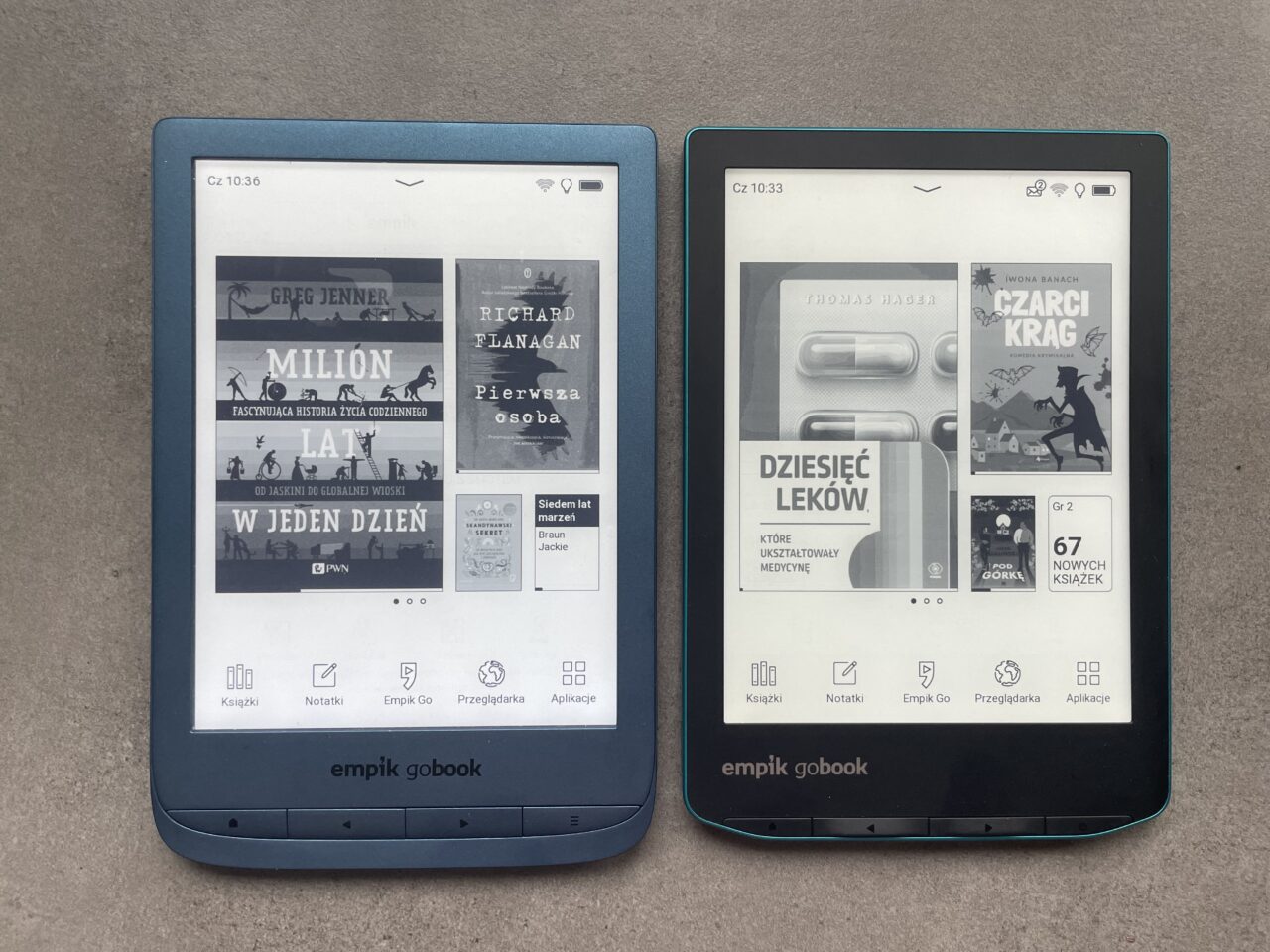 Dwa e-czytniki Empik GoBook z widocznymi na ekranach okładkami e-książek w języku polskim, jeden w niebieskiej obudowie i drugi w czarnej, leżące na szarym tle.