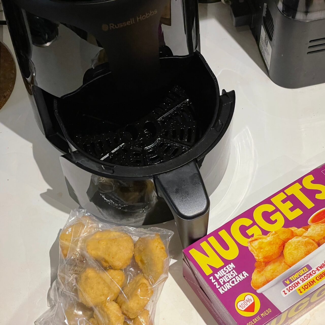 Czarny ekspres do kawy marki Russell Hobbs z otwartą komorą na filtr, obok opakowanie nuggetsów z kurczaka oraz plastikowy worek z widocznymi kilkoma nuggetsami.