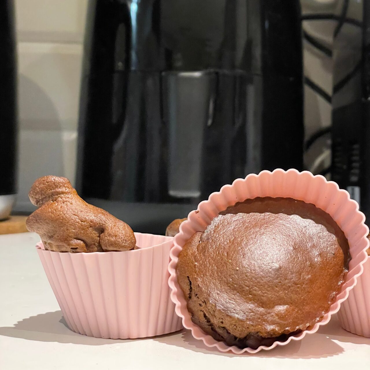 Ciasto w kształcie dinozaura w różowej foremce do muffinek i okrągły czekoladowy babeczek również w różowej foremce, umieszczone na blacie kuchennym na tle czarnej frytkownicy