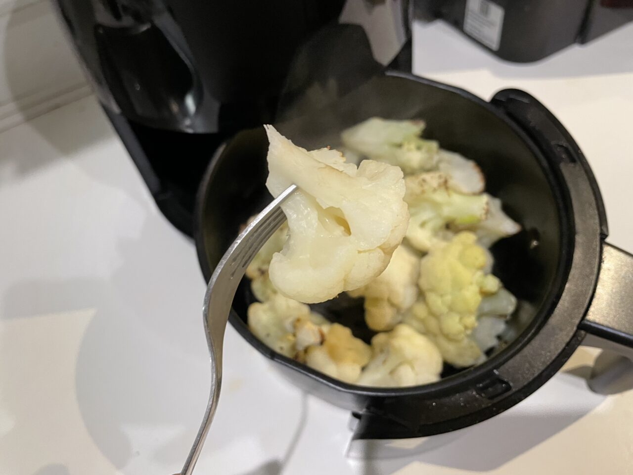 Kawałek gotowanego kalafiora na widelcu nad czarnym garnkiem z resztą warzyw.