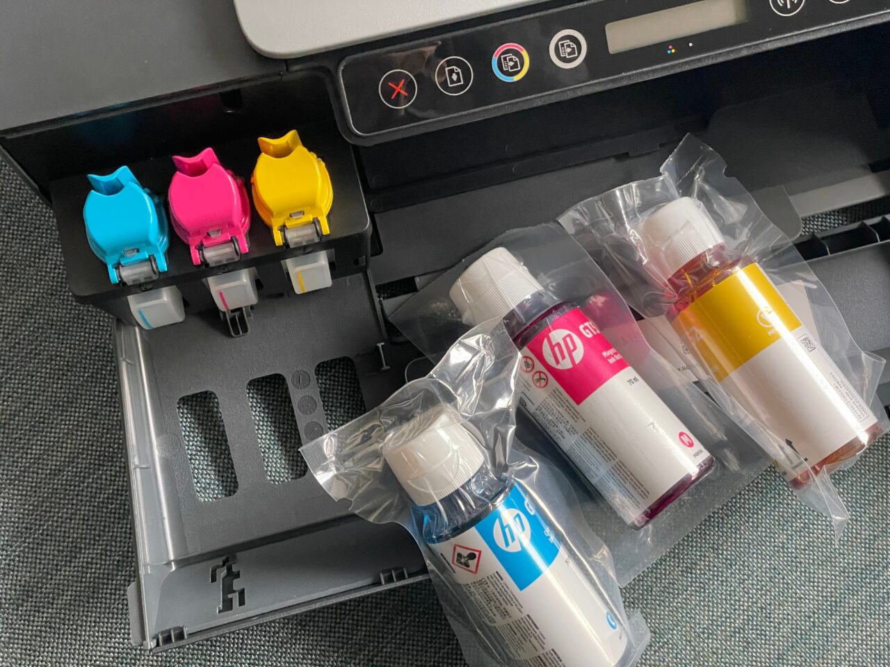Kolorowa drukarka z otwartym przedziałem na wkłady atramentowe i czterema butelkami z atramentem w kolorach niebieskim, różowym, żółtym i czarnym leżącymi obok na blacie.
