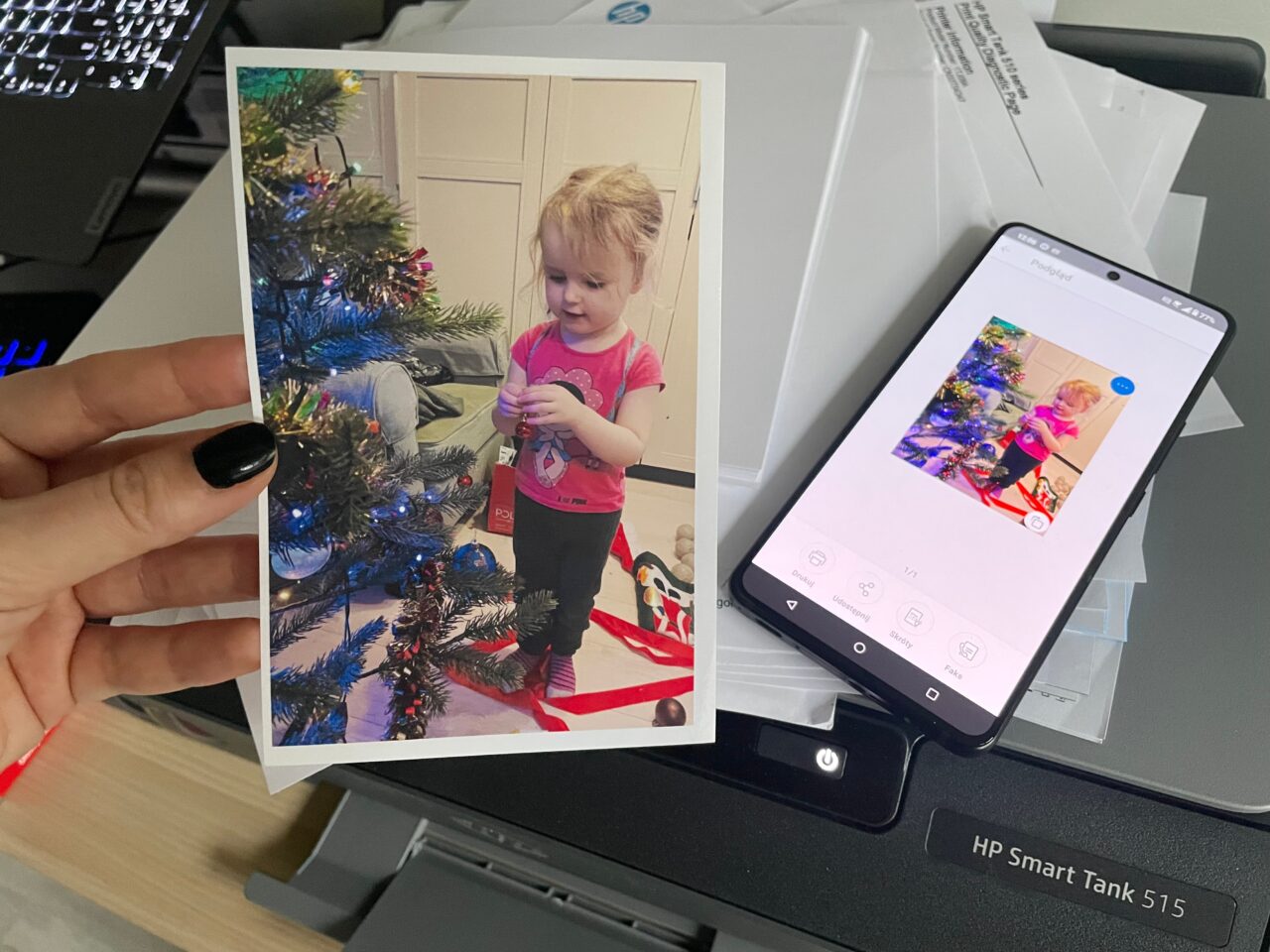 Mała dziewczynka ubierająca choinkę świąteczną na fotografii trzymanej nad drukarką, z odpowiadającym jej obrazem wyświetlanym na ekranie smartfona.