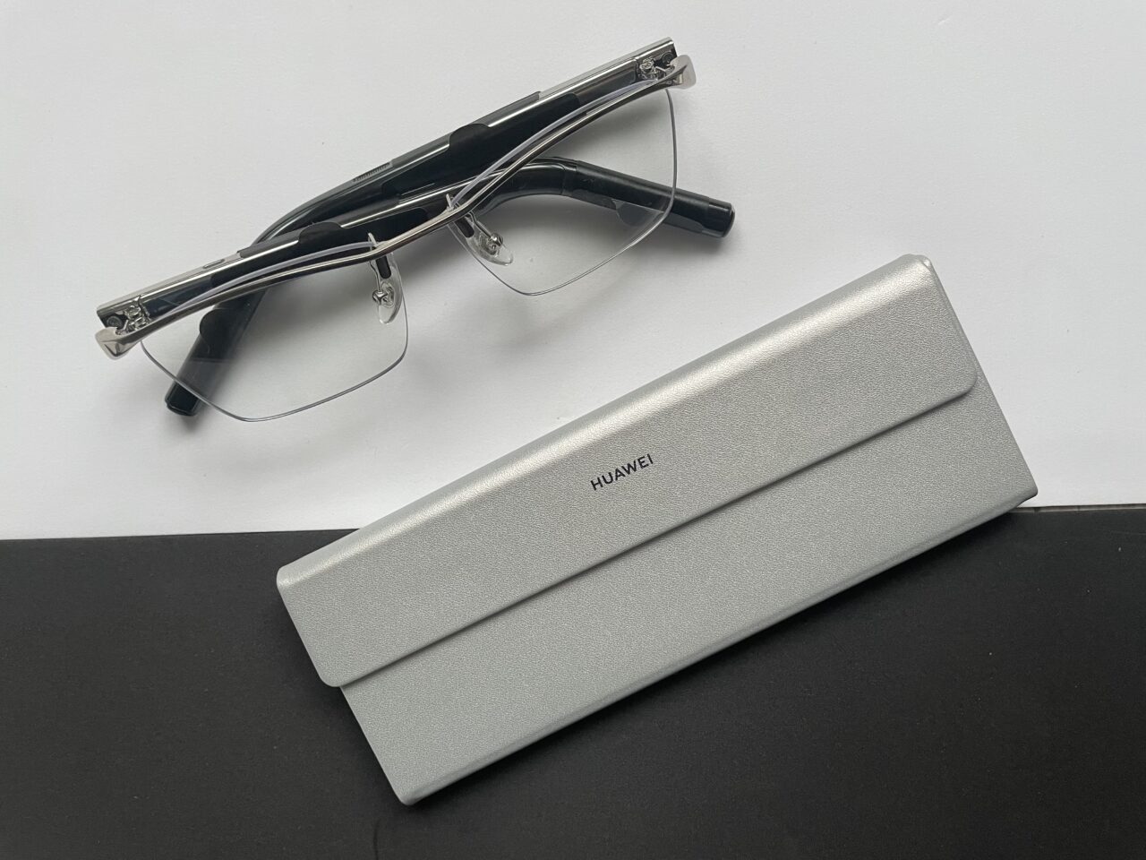 Okulary bezramkowe położone obok metalowego etui z logo Huawei, na białym i czarnym tle.