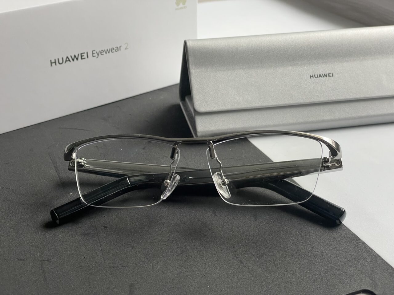 Okulary HUAWEI Eyewear 2 bez oprawek leżą na czarnym podłożu przed otwartym srebrnym etui i białym pudełkiem z logo HUAWEI.