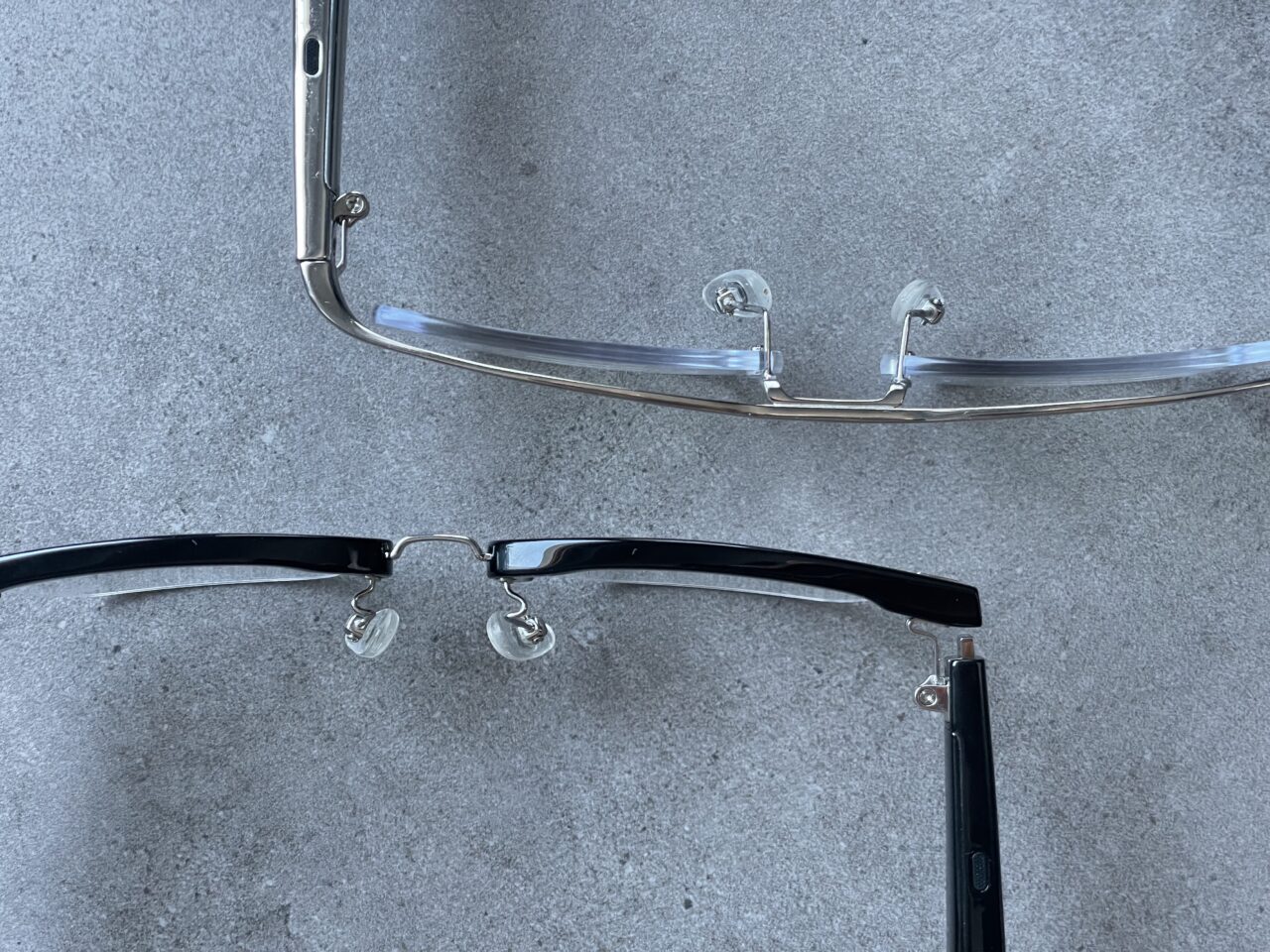 Dwa pary okularów bez soczewek, leżące na szarym betonowym podłożu – jedna para z przezroczystymi, a druga z czarnymi oprawkami.